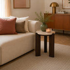 **Round side table with a cream marble top and chunky dark brown timber legs styled beside a cream sofa, decorated with a green vase and lit candle in a warm contemporary living room.**
