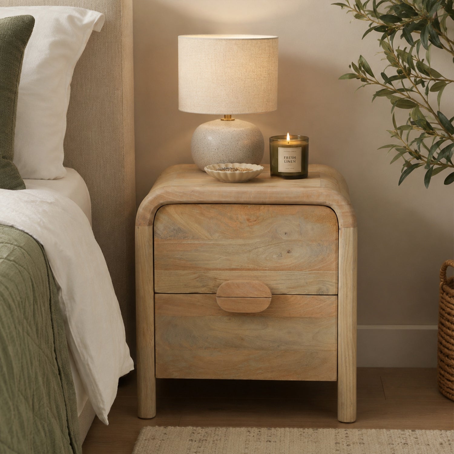 Mango Shona bedside table in natural mango wood with two drawers, styled beside a bed with a ceramic table lamp, candle, decorative dish, and leafy plant in a warm contemporary bedroom.