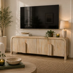 Natural mango wood Shona entertainment unit with rounded edges and sculpted door handles, styled beneath a wall-mounted TV with a table lamp, books, candle, potted plant, and contemporary living room décor.