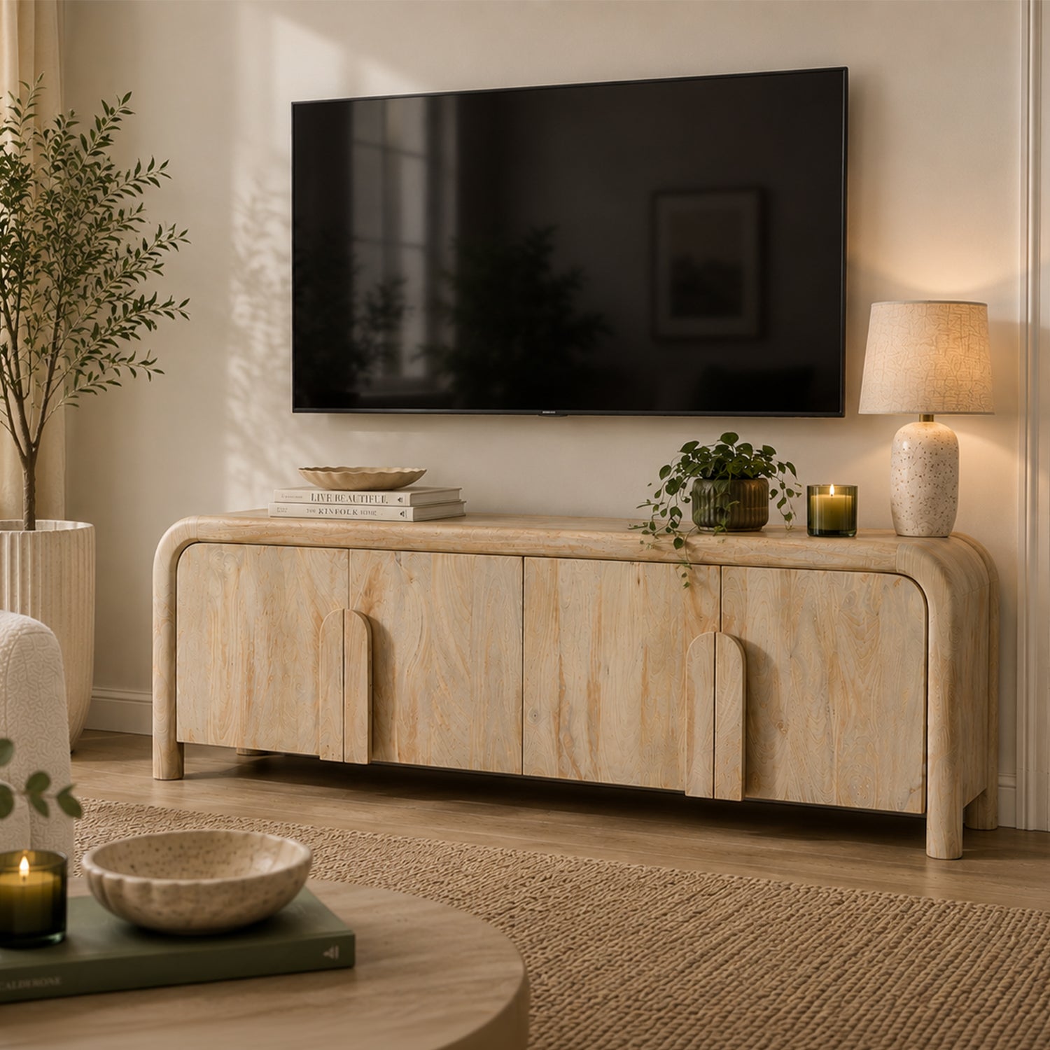 Natural mango wood Shona entertainment unit with rounded edges and sculpted door handles, styled beneath a wall-mounted TV with a table lamp, books, candle, potted plant, and contemporary living room décor.