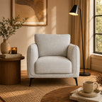 A light gray upholstered armchair with black legs sits in a warm, sunlit living room beside a window, floor lamp, and wooden side table.
