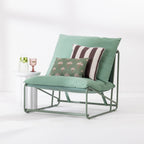 A green outdoor lounge chair with a metal frame, styled with a large green back cushion, a brown and white striped accent pillow, and a smaller green palm-patterned throw pillow. The chair is set against a clean white background next to a white marble side table holding a glass of water and a pink drink.