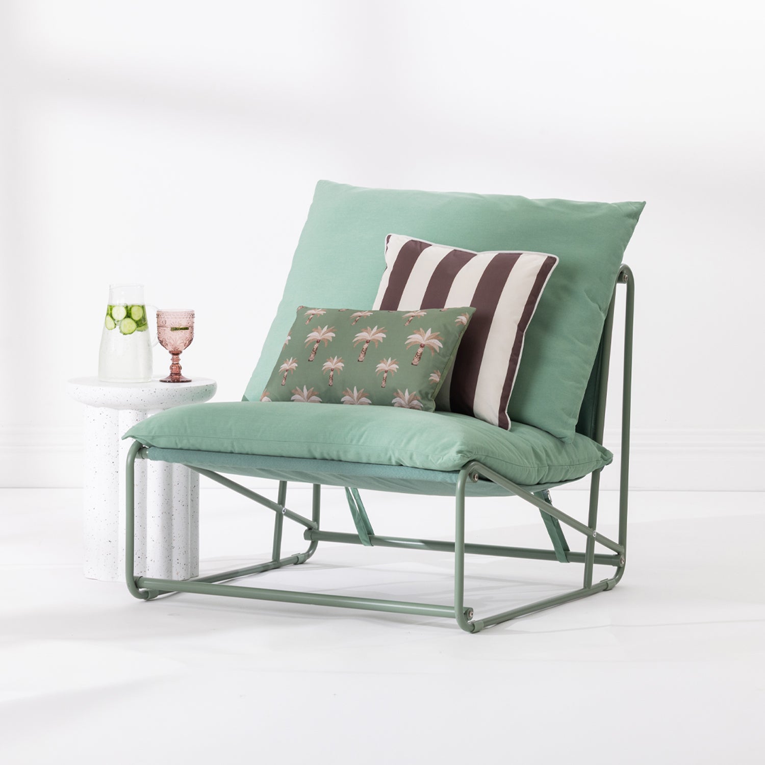 A green outdoor lounge chair with a metal frame, styled with a large green back cushion, a brown and white striped accent pillow, and a smaller green palm-patterned throw pillow. The chair is set against a clean white background next to a white marble side table holding a glass of water and a pink drink.