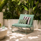 A green outdoor lounge chair with a metal frame, styled with two decorative throw pillows in a tropical patio setting with lush green plants.