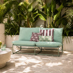 A green outdoor 2 seater sofa with a matching metal frame, styled with three decorative throw pillows on a stone patio in front of a backdrop of large green tropical plants.