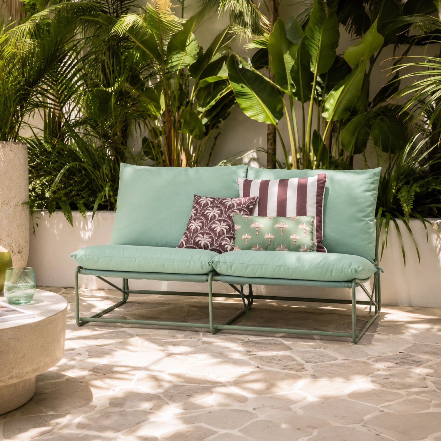 A green outdoor 2 seater sofa with a matching metal frame, styled with three decorative throw pillows on a stone patio in front of a backdrop of large green tropical plants.
