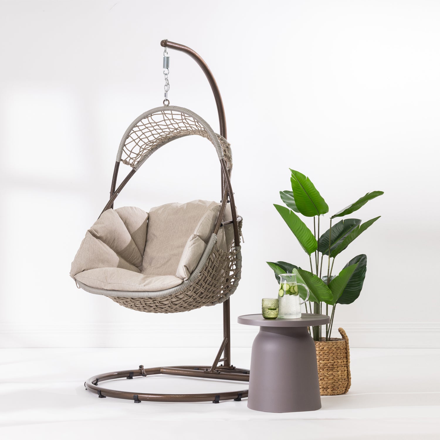 A hanging egg chair with a woven wicker body, beige cushions, and a dark metal stand, photographed in a bright white studio setting.