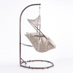 A hanging egg chair with a woven wicker body, beige cushions, and a dark metal stand, photographed in a bright white studio setting.