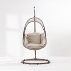 A hanging egg chair with a woven wicker body, beige cushions, and a dark metal stand, photographed in a bright white studio setting.