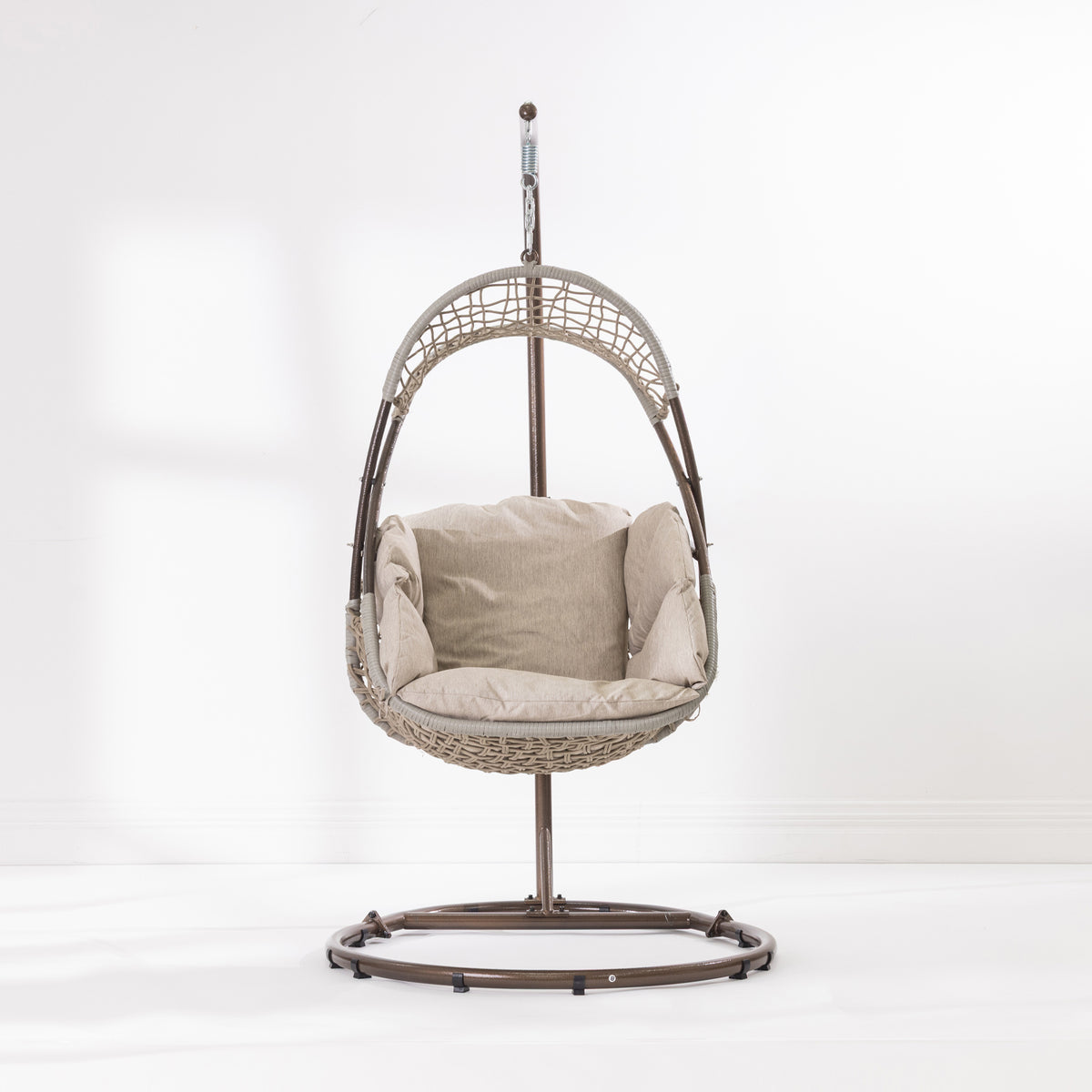 A hanging egg chair with a woven wicker body, beige cushions, and a dark metal stand, photographed in a bright white studio setting.