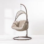 A hanging egg chair with a woven wicker body, beige cushions, and a dark metal stand, photographed in a bright white studio setting.