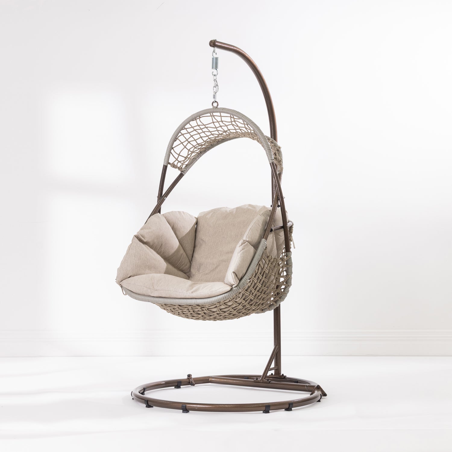 A hanging egg chair with a woven wicker body, beige cushions, and a dark metal stand, photographed in a bright white studio setting.