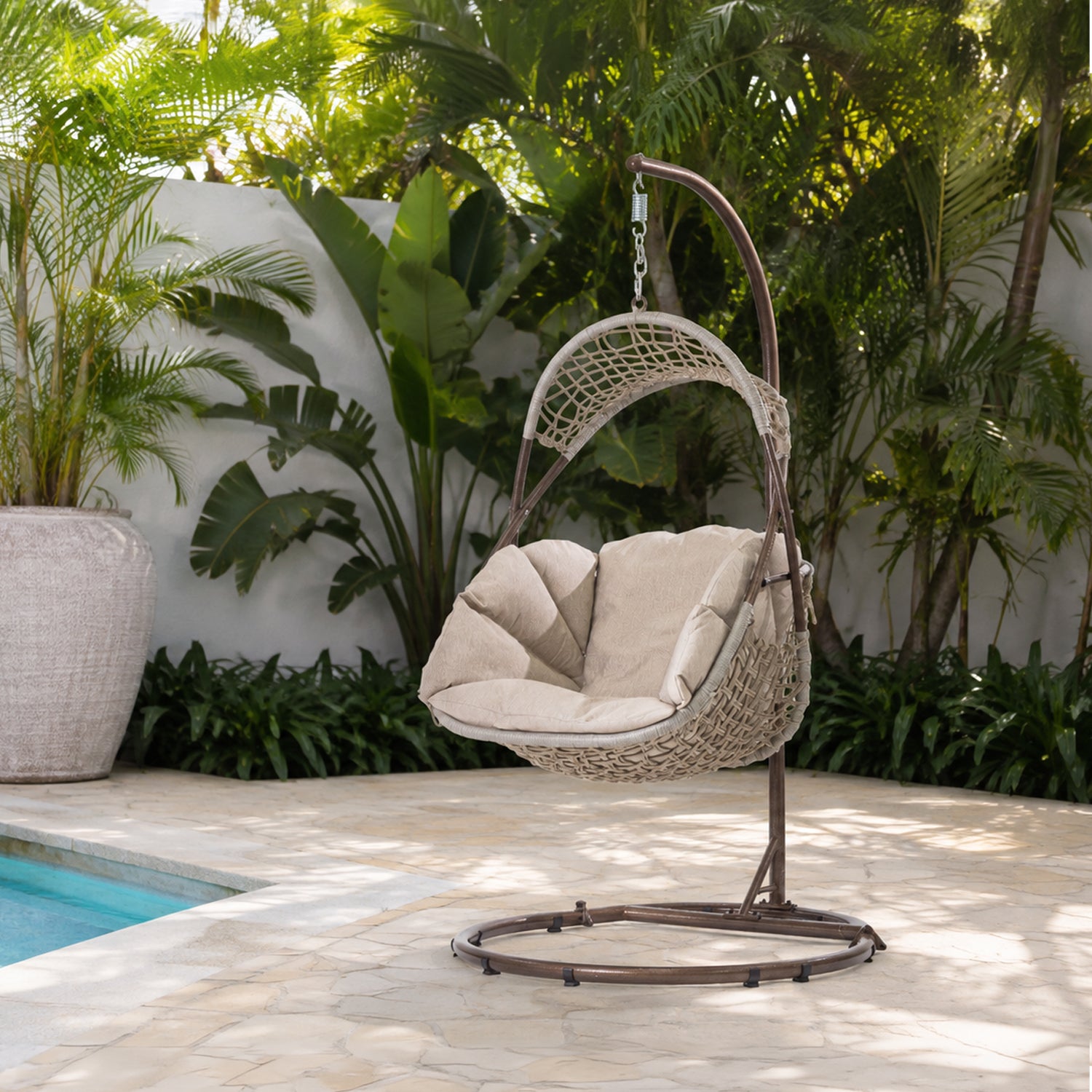 A woven hanging egg chair with a natural cushion sits on a metal stand in a tropical backyard patio setting next to a swimming pool.