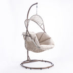 A hanging egg chair with a woven wicker body, beige cushions, and a dark metal stand, photographed in a bright white studio setting.