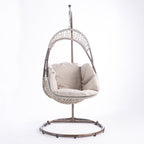 A hanging egg chair with a woven wicker body, beige cushions, and a dark metal stand, photographed in a bright white studio setting.
