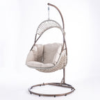 A hanging egg chair with a woven wicker body, beige cushions, and a dark metal stand, photographed in a bright white studio setting.
