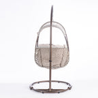 A hanging egg chair with a woven wicker body, beige cushions, and a dark metal stand, photographed in a bright white studio setting.