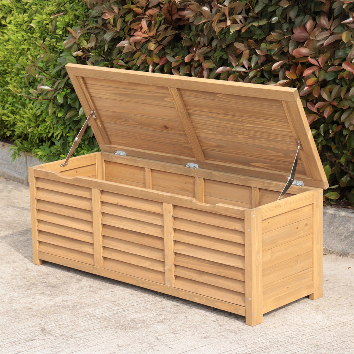 A natural wood outdoor storage box with a slatted front and an open hinged lid supported by metal struts, shown on pavement with green foliage in the background.