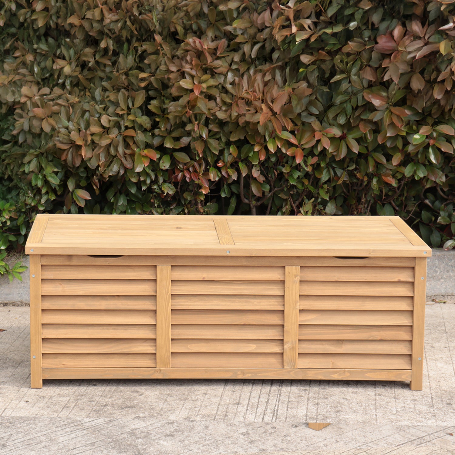 A rectangular wooden storage bench sits outdoors on a concrete surface in front of a dense green and reddish-brown hedge.
