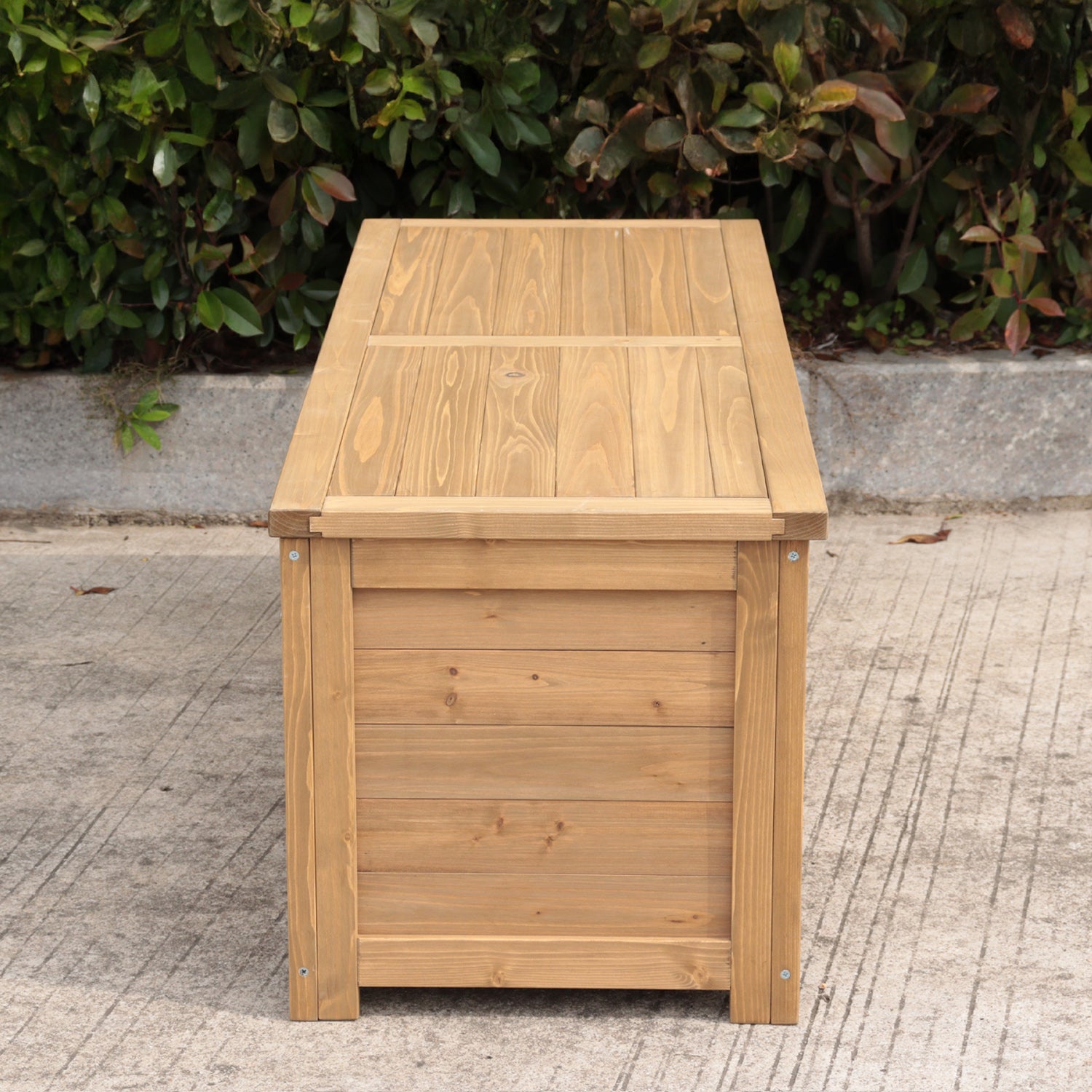 A rectangular wooden storage box made of light natural wood, photographed outdoors on a concrete surface with green foliage in the background.