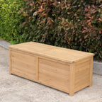 A rectangular wooden storage box with a light natural wood finish, positioned outdoors on a concrete surface in front of a green and reddish leafy hedge.