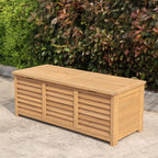 A rectangular wooden storage bench sits outdoors on a concrete surface in front of a dense green and reddish-brown hedge.