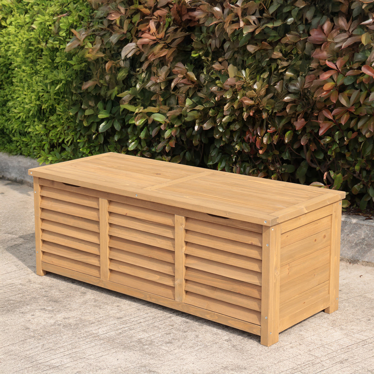 A rectangular wooden storage bench sits outdoors on a concrete surface in front of a dense green and reddish-brown hedge.