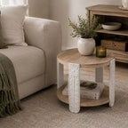 Round natural wood side table with carved antique white legs styled beside a cream sofa with a ceramic vase, greenery and candle in a warm neutral living room.