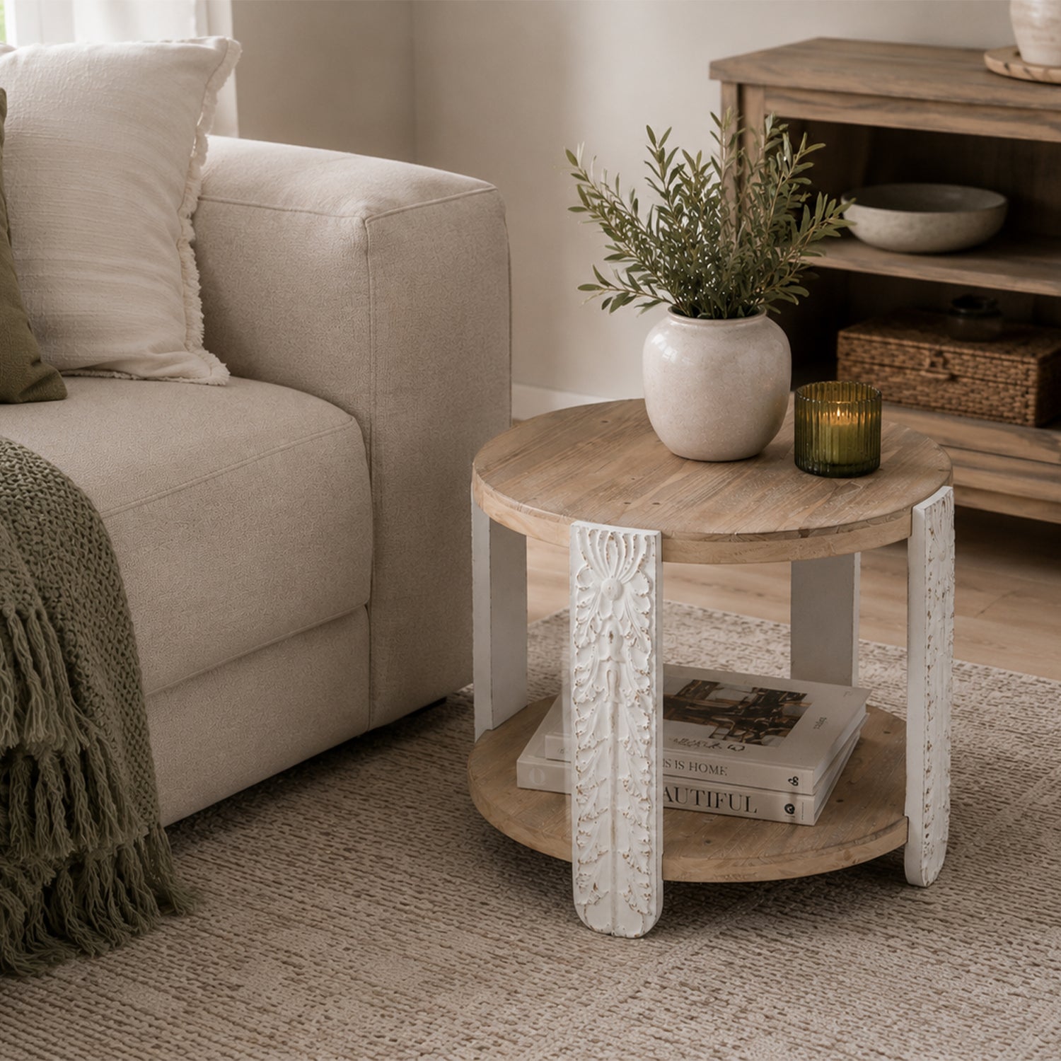 Round natural wood side table with carved antique white legs styled beside a cream sofa with a ceramic vase, greenery and candle in a warm neutral living room.