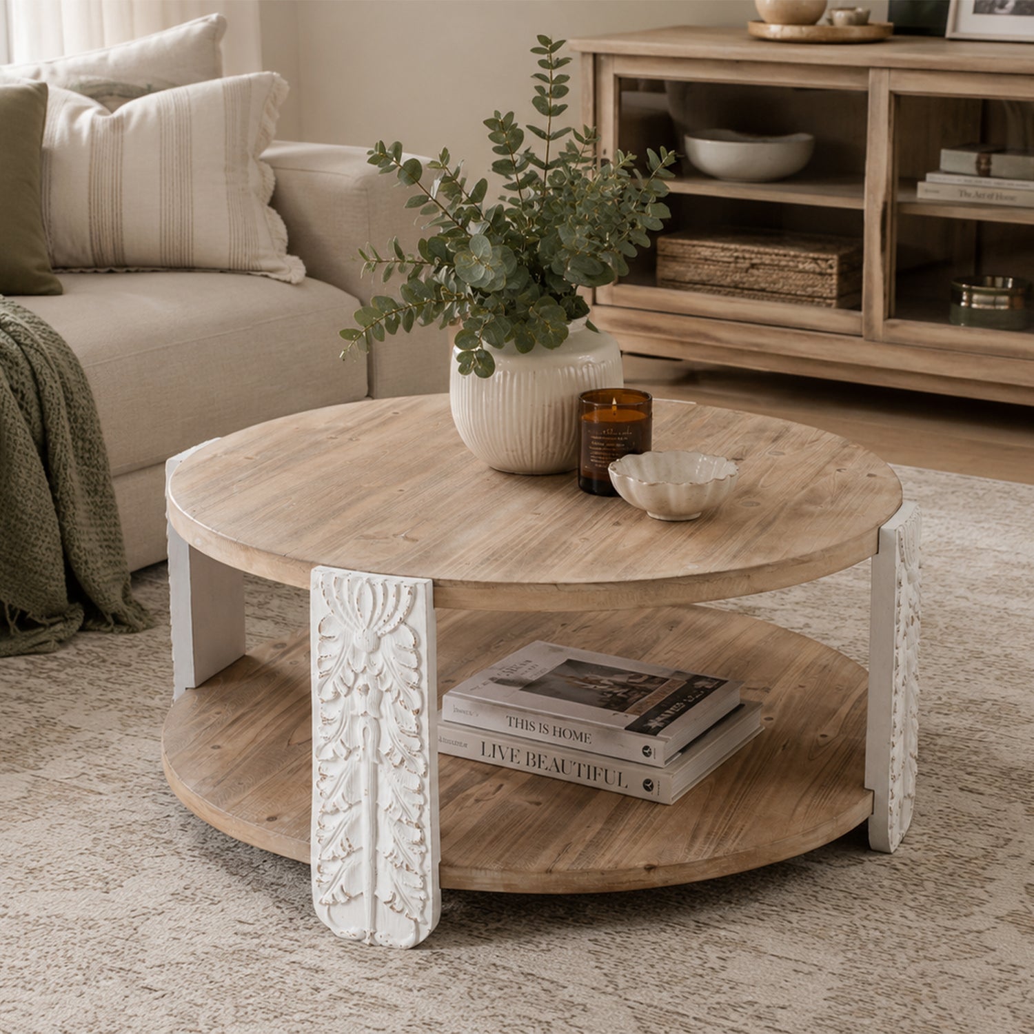 Round natural wood coffee table with a lower shelf and carved antique white legs featuring decorative floral detailing.