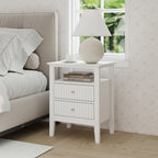 A white two-drawer bedside table with an open shelf, topped with a lamp, books, and a cup, sits beside an upholstered bed in a bright, softly decorated bedroom.