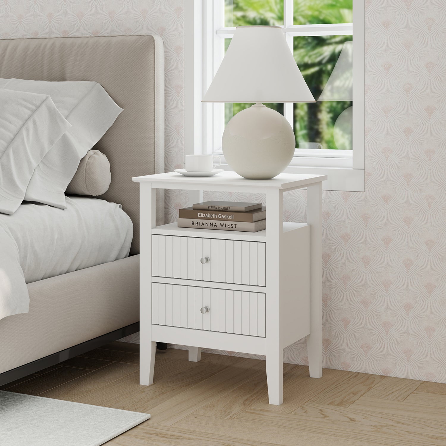 A white two-drawer bedside table with an open shelf, topped with a lamp, books, and a cup, sits beside an upholstered bed in a bright, softly decorated bedroom.