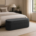 Black pleated velvet storage bench positioned at the foot of a bed in a contemporary bedroom, featuring a softly upholstered lid, vertical channel-stitched detailing, a neutral rug, black bedside table, and warm natural light.