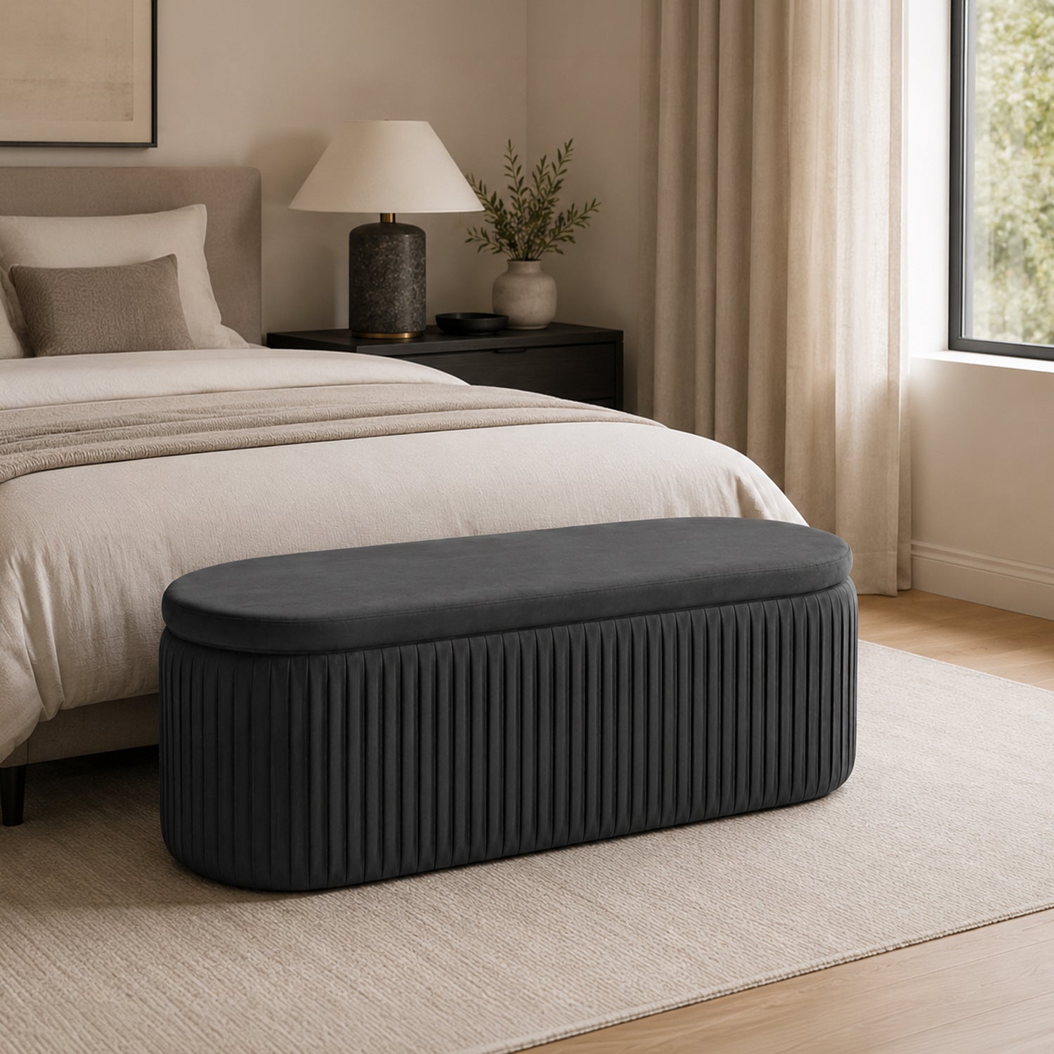 Black pleated velvet storage bench positioned at the foot of a bed in a contemporary bedroom, featuring a softly upholstered lid, vertical channel-stitched detailing, a neutral rug, black bedside table, and warm natural light.