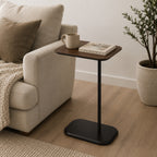 A walnut-look side table with a slim black metal base is styled beside a sofa with a mug and book, creating a warm, modern accent for relaxed everyday living.