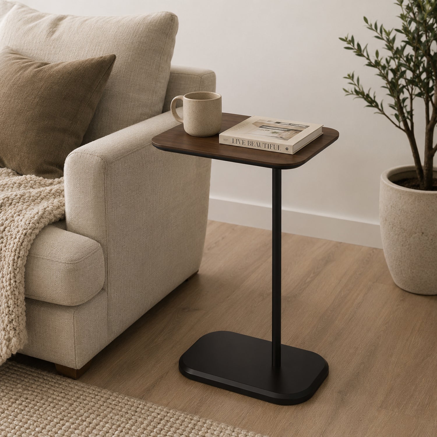 A walnut-look side table with a slim black metal base is styled beside a sofa with a mug and book, creating a warm, modern accent for relaxed everyday living.