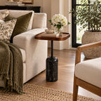 A modern side table with a rounded wooden top, a black marble base, and a single black metal stem, styled in a cozy living room next to a cream sofa.