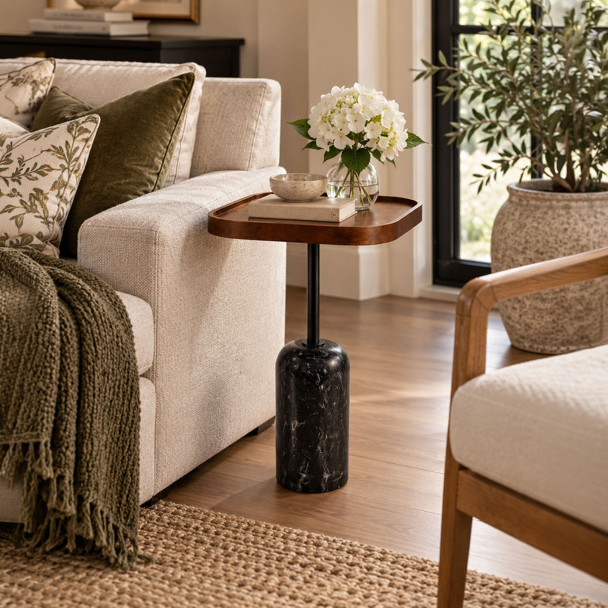 A modern side table with a rounded wooden top, a black marble base, and a single black metal stem, styled in a cozy living room next to a cream sofa.