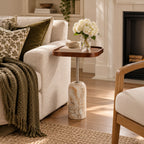 A small round side table with a dark wood top and a marble-look pedestal base in a warm white tone, styled in a cozy living room next to a cream sofa with a green throw blanket and white flowers on top.