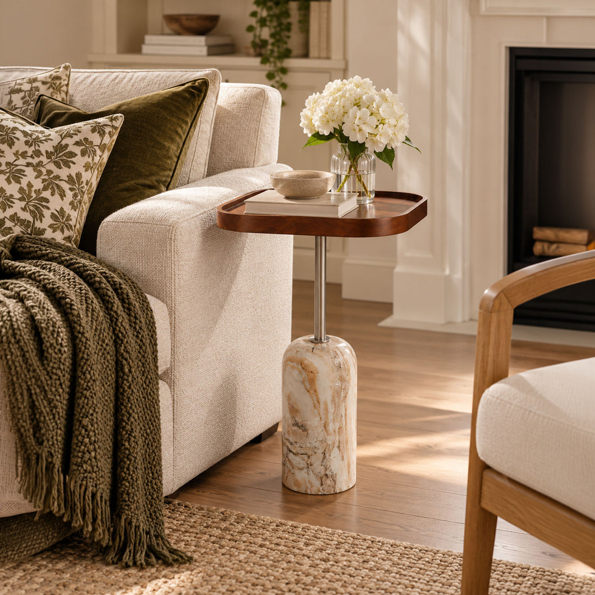 A small round side table with a dark wood top and a marble-look pedestal base in a warm white tone, styled in a cozy living room next to a cream sofa with a green throw blanket and white flowers on top.