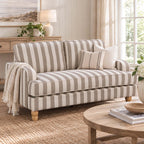 A striped loveseat sofa in beige and off-white with wooden legs, styled in a bright, coastal-themed living room with a throw blanket, pillows, a wooden coffee table, and a window.