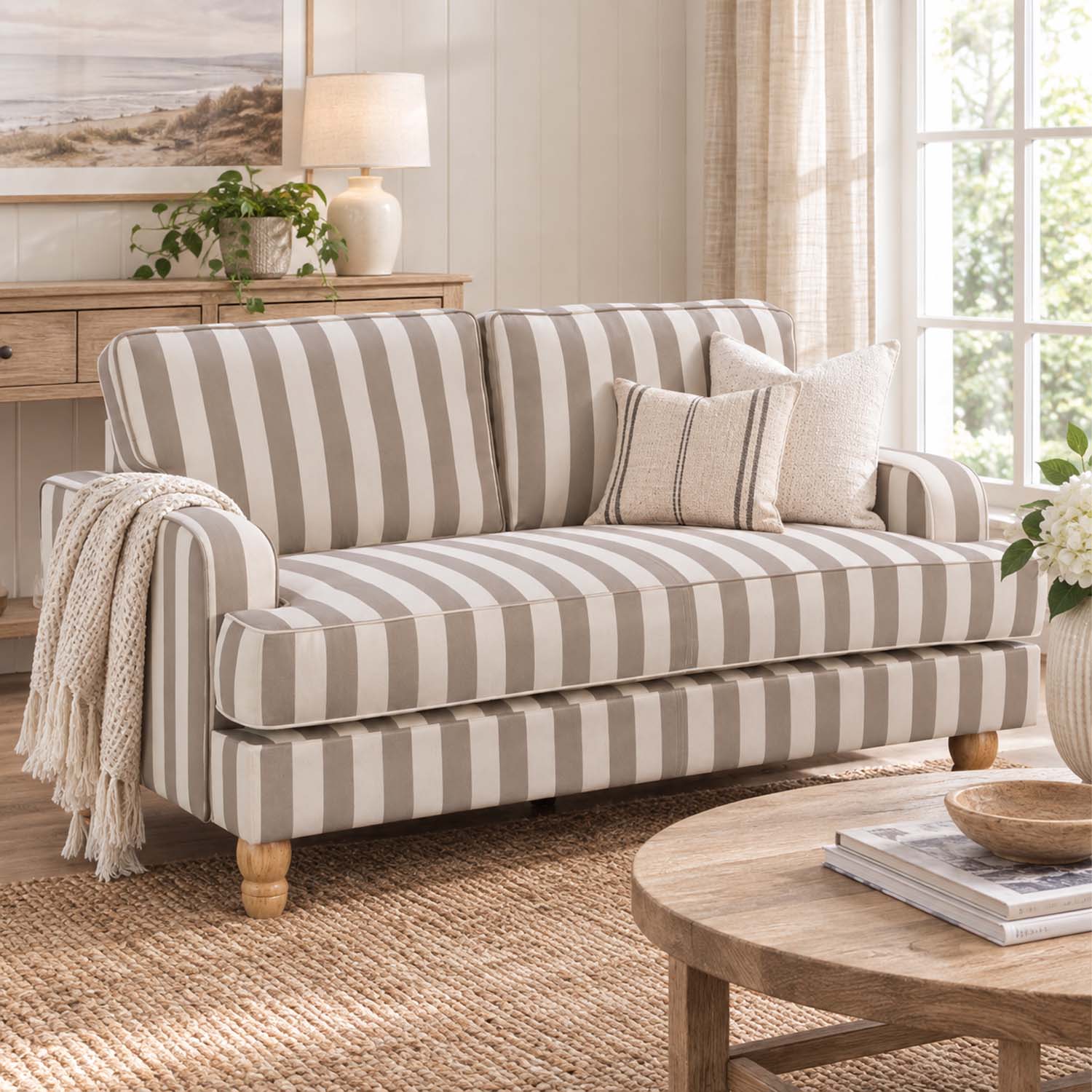 A striped loveseat sofa in beige and off-white with wooden legs, styled in a bright, coastal-themed living room with a throw blanket, pillows, a wooden coffee table, and a window.