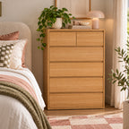 Home Co. Olsen 6 Drawer Tallboy in Natural styled in a warm bedroom setting beside a bed, with a lamp, books and trailing greenery displayed on top.