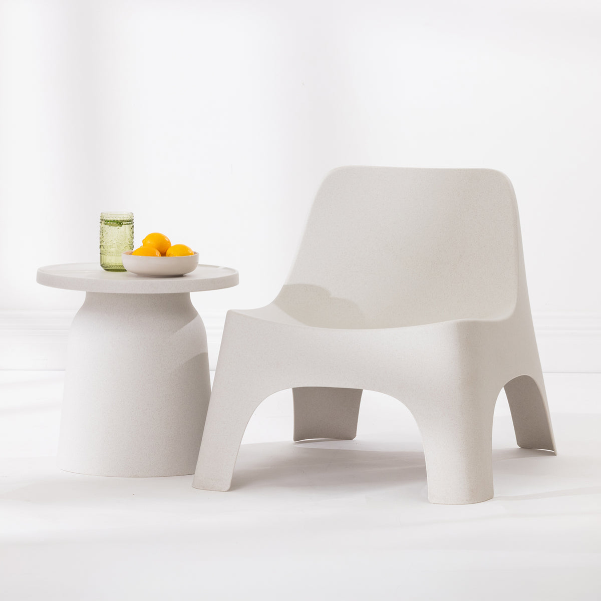 A modern moulded plastic lounge chair in a cream colour, shown next to a matching side table with a glass and yellow fruit on a clean white studio background.