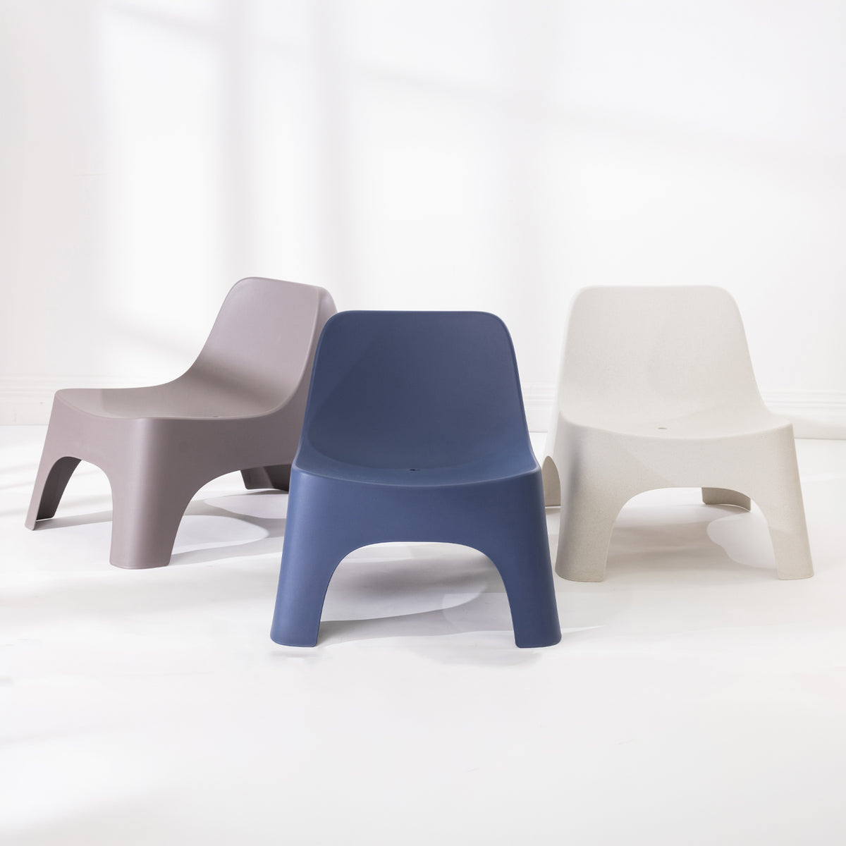 Three moulded plastic chairs in neutral colours: a mocha on the left, a navy blue in the centre, and an cream on the right, arranged on a white floor against a white wall.