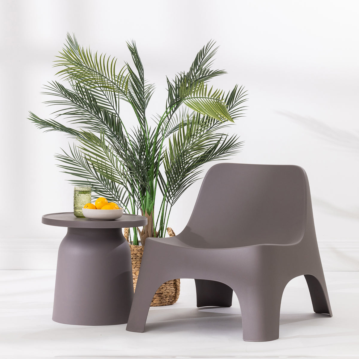 A modern moulded plastic lounge chair in mocha, styled next to a matching side table with a bowl of oranges and a potted palm plant on a white floor against a white wall.