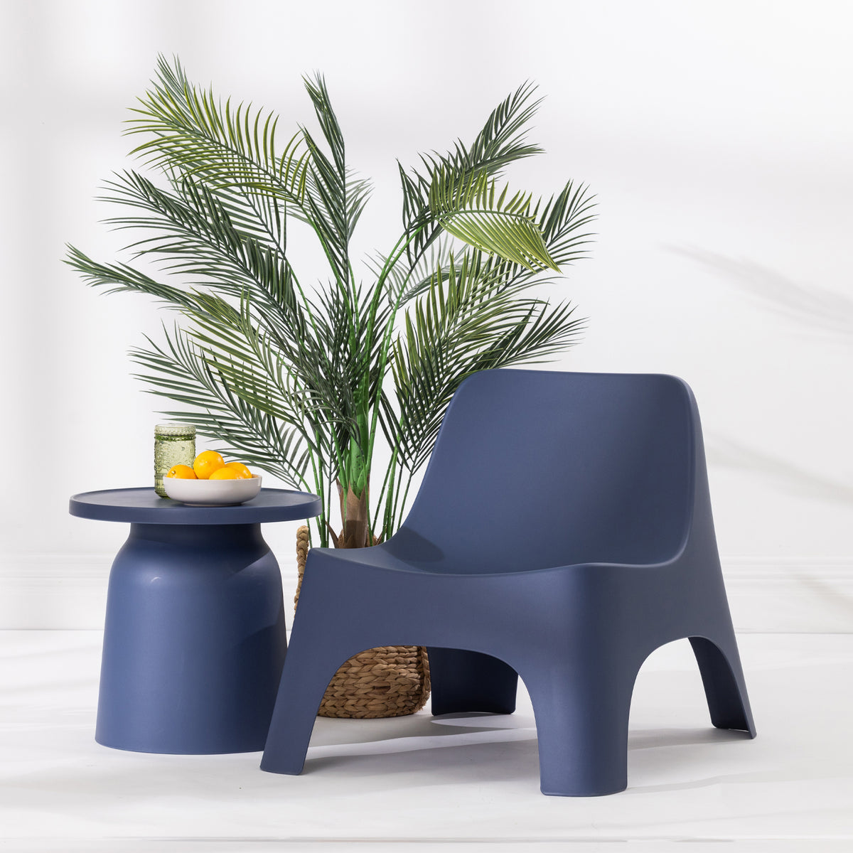 A modern moulded plastic lounge chair in navy blue, styled next to a matching side table with a bowl of oranges and a potted palm plant on a white floor against a white wall.