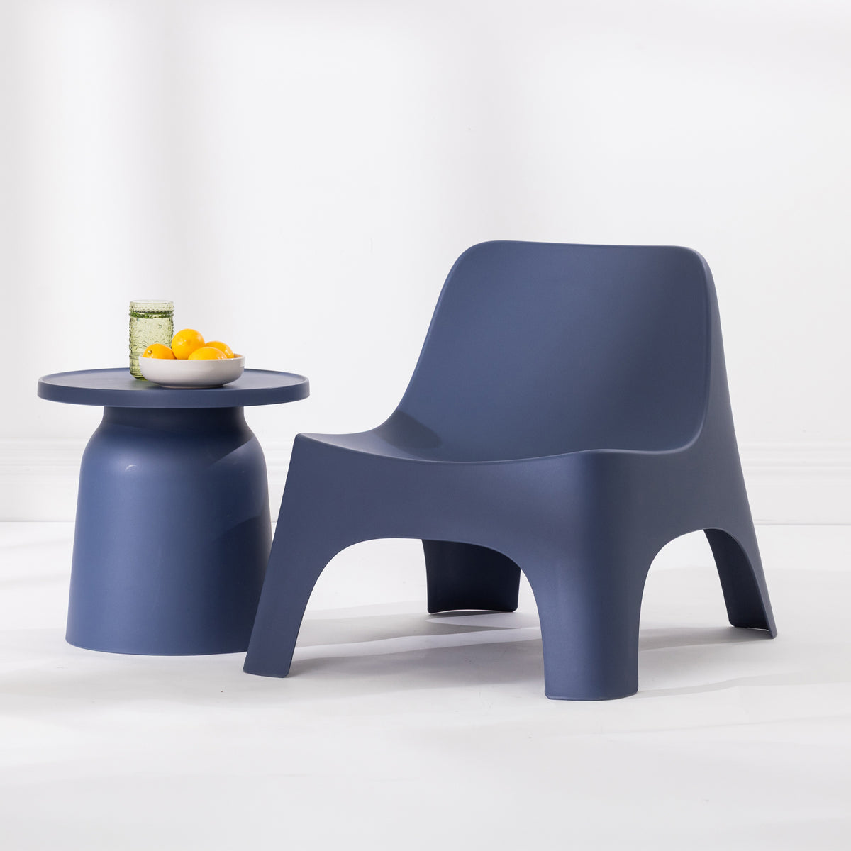 A modern moulded plastic lounge chair in a navy blue colour, shown next to a matching side table with a glass and yellow fruit on a clean white studio background.