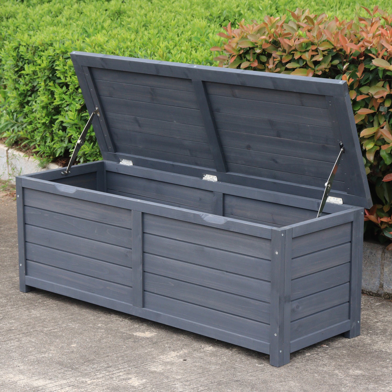 A charcoal wooden storage box with its lid propped open using two metal support rods, set outdoors on a concrete surface with green bushes in the background.