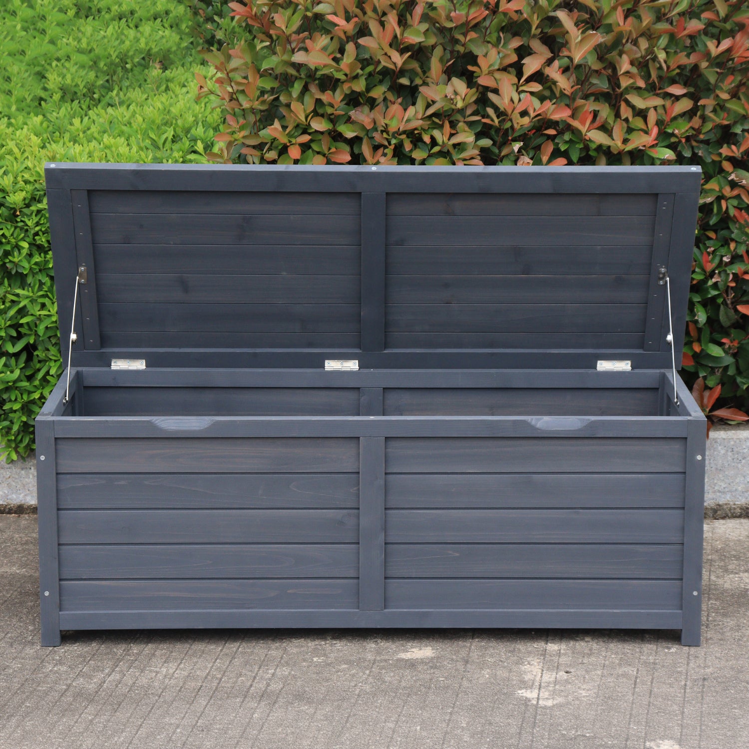 A charcoal wooden storage box with its lid propped open using two metal support rods, set outdoors on a concrete surface with green bushes in the background.